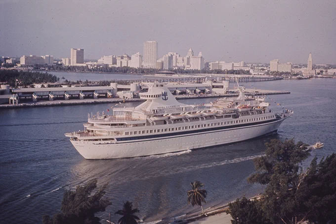 A Royal Caribbean cruise ship navigates a waterway. Miami's skyline is in the background, featuring various buildings and structures. The ship is white with several decks and the 'ROYAL CARIBBEAN' logo.