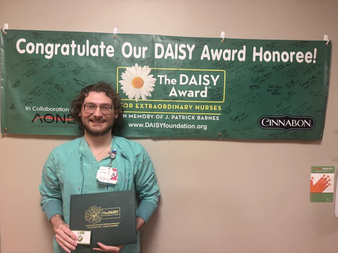 A person stands smiling in front of a green banner that reads, "Congratulations Our DAISY Award Honoree!" The DAISY Foundation logo and Cinnabon logo are visible. The person is holding a certificate folder labeled "The DAISY Award for Extraordinary Nurses" with a badge clipped to their clothing.
