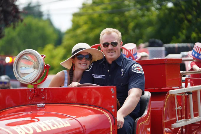 A firefighter and a person next to them pose for a picture in a fire truck at the front of a parade.