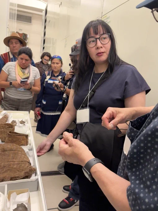 There is a group of people in the National Museum of the American Indian's collections storage observing cultural belongings. One person in the foreground is speaking to someone out of frame. The other group members are taking notes or photos.