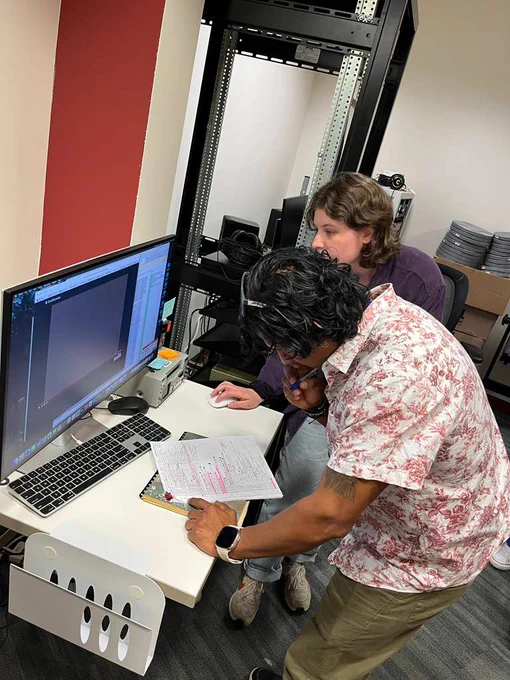 Two people stand at a computer looking at film footage on the monitor. One person is moving the mouse of the computer, while the other is taking notes on a sheet of paper in front of them.