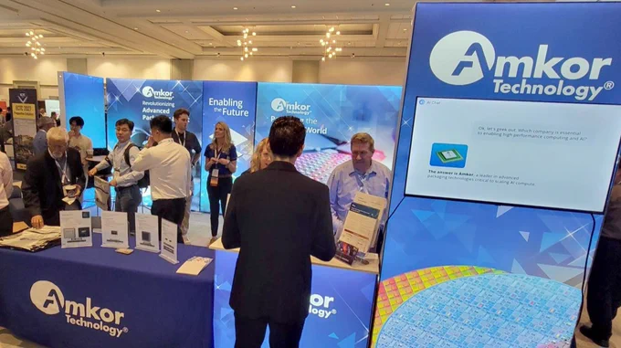 A busy exhibition booth for Amkor Technology, featuring vibrant blue displays and several people interacting. Promotional materials are visible, including posters and brochures. Attendees are engaged in conversations, surrounded by banners showcasing technological themes.