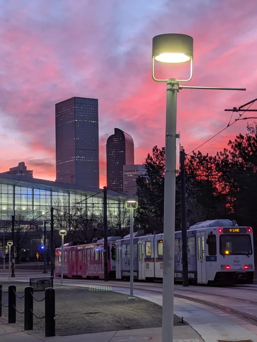 The RTD H-line light rail train passes by the Colorado Convention Center next to a line of street lamps. Behind it is a pink sunrise and some of Denver's skyline, including the famous cash register building.