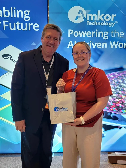 Two people are smiling while standing in front of a backdrop featuring the Amkor Technology logo and the phrases "Enabling the Future" and "Powering the AI-Driven World." One person is holding a white Amkor-branded gift bag.