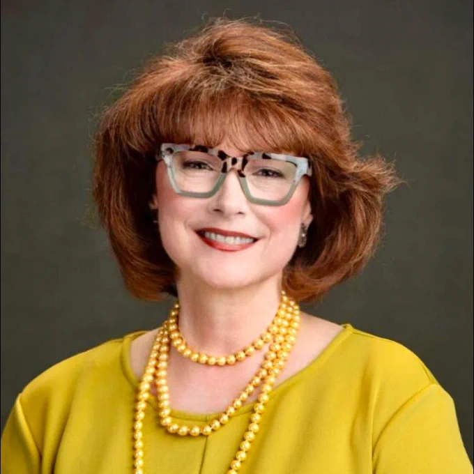 Head shot Barbara Chapman, smiling, wearing glasses and a bright gold sweater.  Black background.