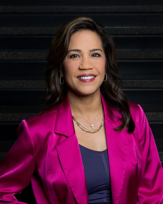 Head shot Kate Taylor, smiling, wearing bright pink blazers, black undershirt.  Black background.