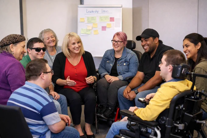 A diverse group of people sitting in a circle engaged in a discussion with a whiteboard labeled 'Leadership Ideas' in the background.
