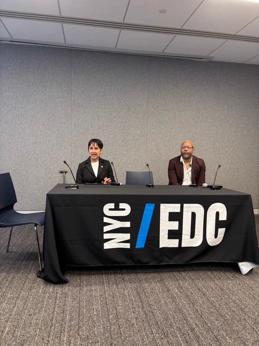 Commissioner Agarwal and James Irvin representing the NYC Mayor’s Office for People with Disabilities are seated at a table covered with a black tablecloth displaying the white and blue “NYC EDC” logo. Each has a microphone in front of them, suggesting a panel discussion or press conference. The setting is a professional conference room with gray walls and carpeted flooring, conveying a formal and organized atmosphere.
