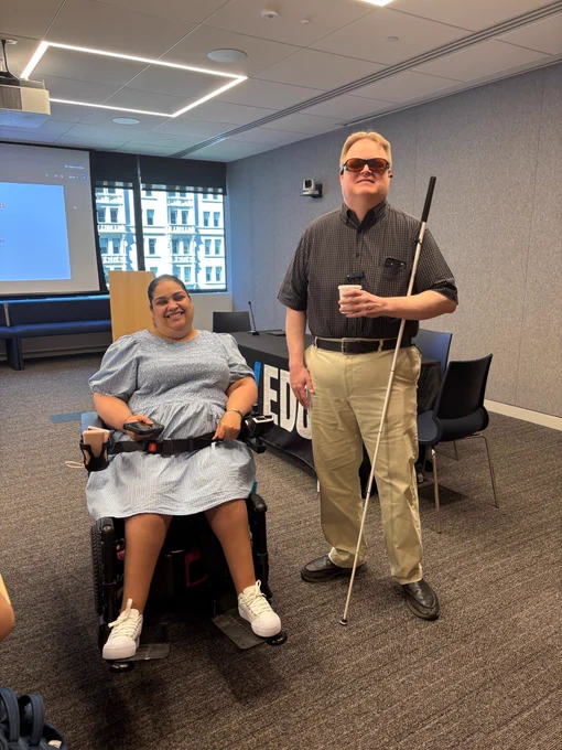 Arthur Jacobs, wearing dark glasses and holding a white cane, stands beside Crystal Rivera, who is using a motorized wheelchair. They are positioned in front of a conference room, which features chairs and a presentation screen in the background.