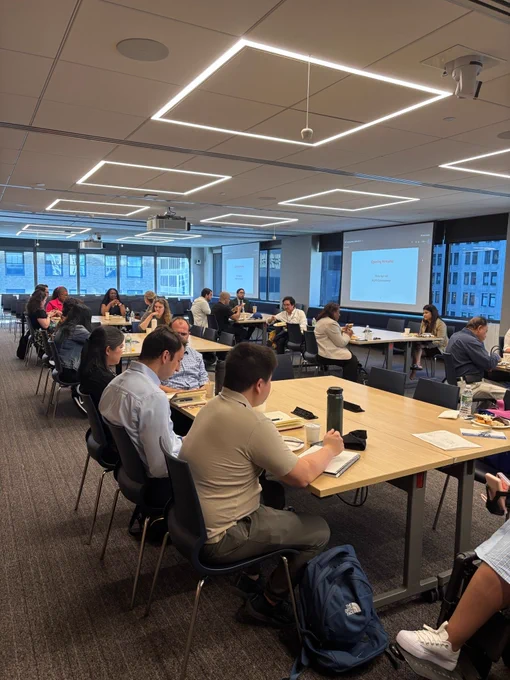 People sit around large rectangular tables in a modern conference room with geometric ceiling lights and tall windows showing city buildings. A presentation slide at the front reads “Opening Remarks” with “Nisha Agarwal Commissioner ” underneath. Participants appear engaged, with laptops, notebooks, and water bottles on the tables.