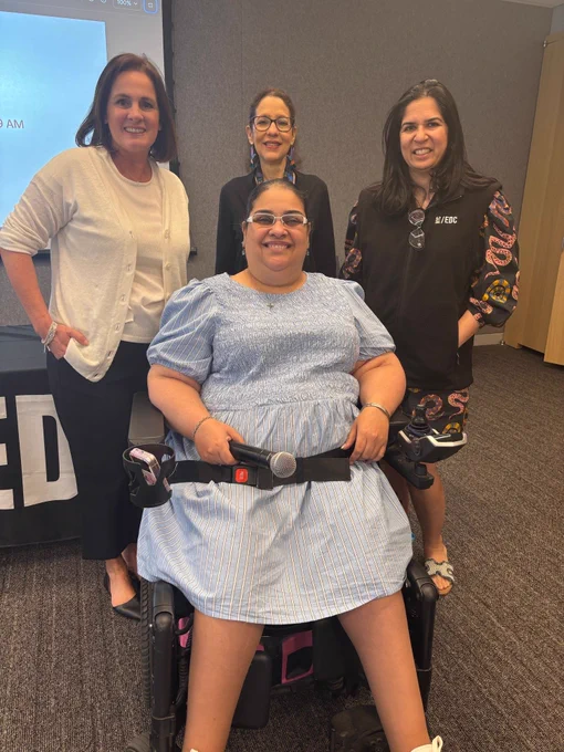 Four people pose together in a conference room. Crystal Rivera, in a motorized wheelchair, sits at the front holding a microphone. Seema Malik (Right), Vanessa Ramos (Center), and Jennifer Lin Perry (Left) are standing behind her. A gray wall, a wooden cabinet, and part of a projected blue slide are visible in the background, suggesting a professional event or presentation.
