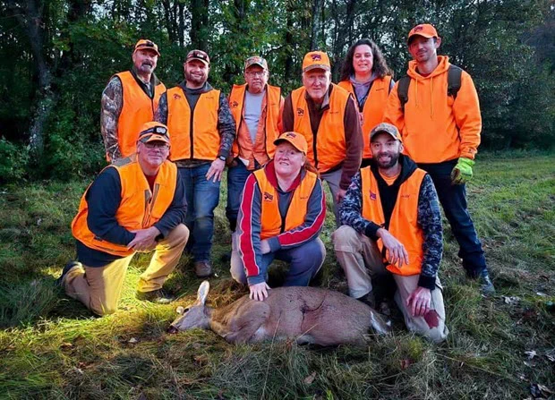 A small group of hunters wearing blaze orange smiles for a photo behind three hunters kneeling next to a deer harvested during a fall hunt.