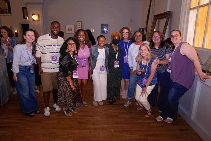 A group of people posing together indoors, all wearing purple lanyards. The room has wooden floors and light-colored walls, with artwork visible in the background. Everyone is smiling.