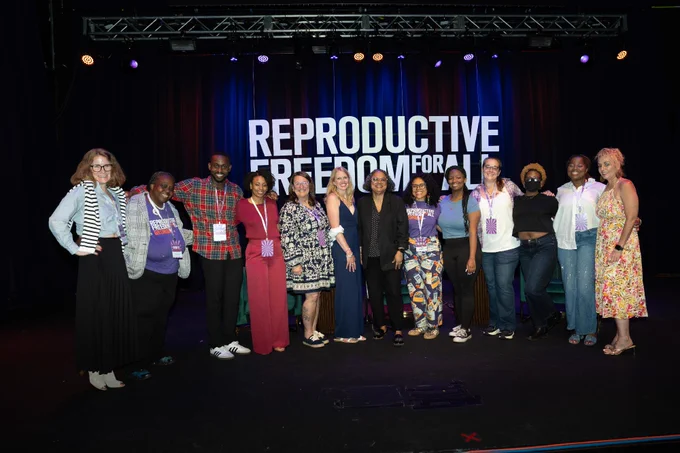 A diverse group of individuals stands on stage in front of a backdrop that reads "Reproductive Freedom for All." They are smiling and dressed casually.
