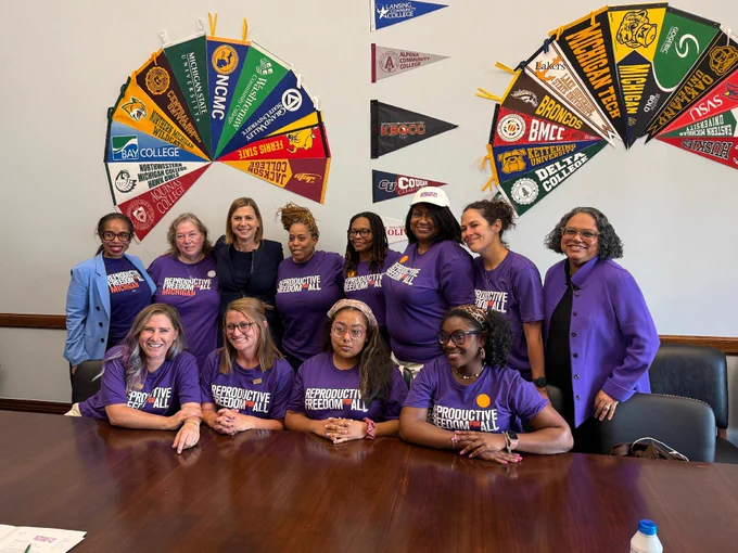 A group of individuals wearing matching purple shirts with the text "Reproductive Freedom For All" are gathered around a conference table. Behind them, a wall displays numerous college pennants, including ones from Fayetteville State University, Morgan State University, and Spelman College.
