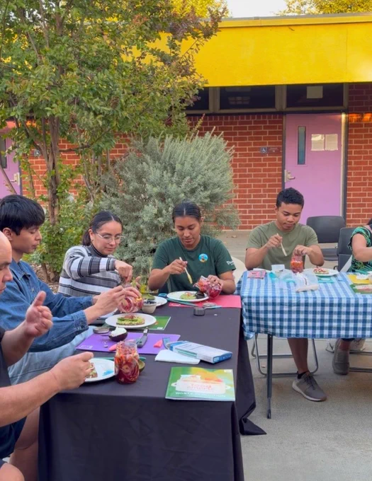 A group of people seated at a table outdoors are engaged in an activity. The table is covered with a black and blue checkered cloth and various craft supplies. In the background, there's a building with a brick wall and purple doors.
