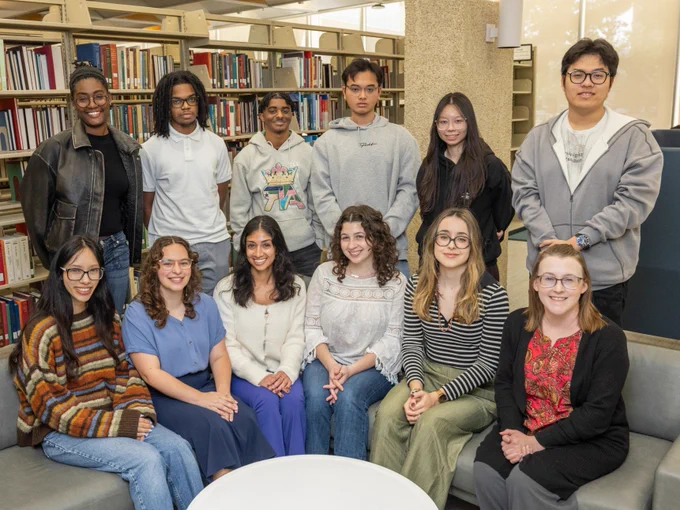 Group of twelve people posing together in a library. They are gathered around a small round table, with some sitting on a sofa and others standing behind. Shelves filled with books are visible in the background.