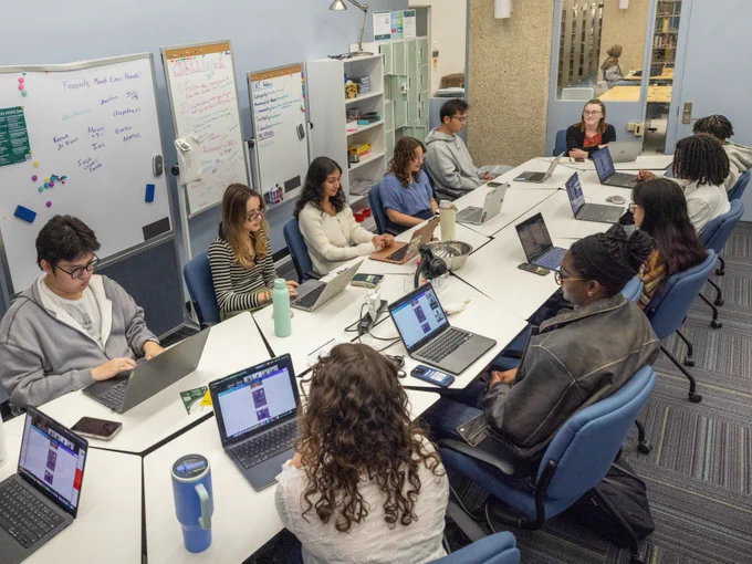 A diverse group of people are sitting around a large, rectangular table, using laptops and writing materials. The room has blue walls with whiteboards filled with notes. Bookshelves and additional seating are in the background. The setting suggests a collaborative or educational environment.