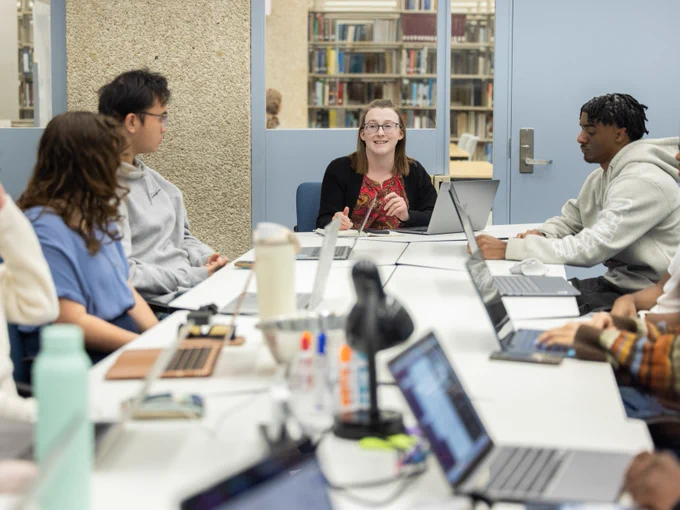 A group of people sitting around a table in a library, engaging in discussion. Laptops, notebooks, and water bottles are placed on the table. Bookshelves are visible in the background.