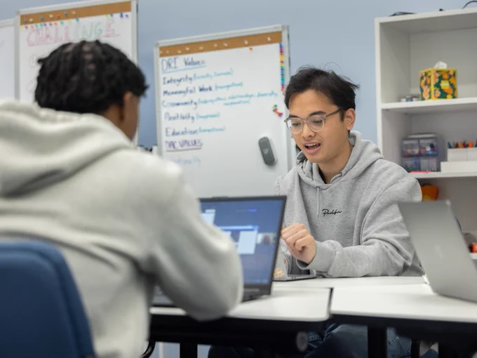 Two people sitting at a table with laptops, engaged in discussion. A whiteboard with notes and a shelf with books and supplies are visible in the background.