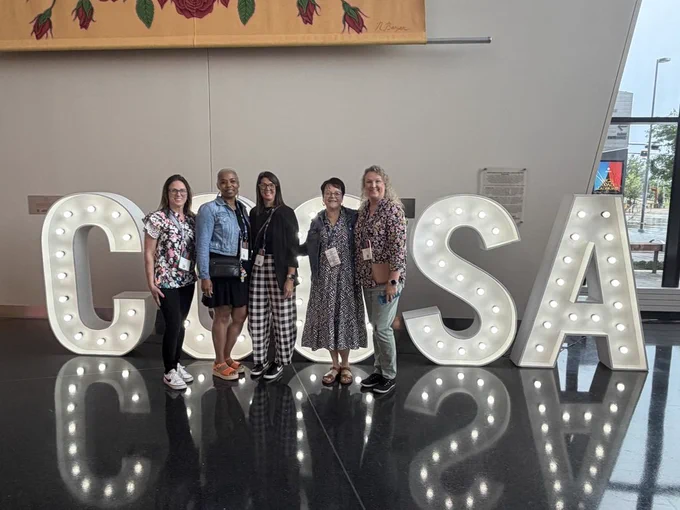 Five women in casual attire standing behind large letters spelling out "CONSA". A decorative wall backdrop behind them.
