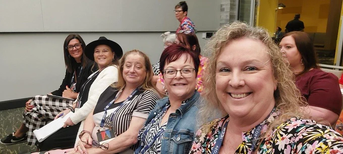 A group of women seated in chairs at an event. They wear different outfits and accessories.