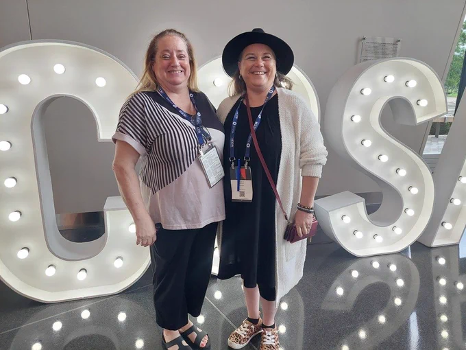Two women in front of a large light display spelling out "CS". One woman wears a hat and sandals.
