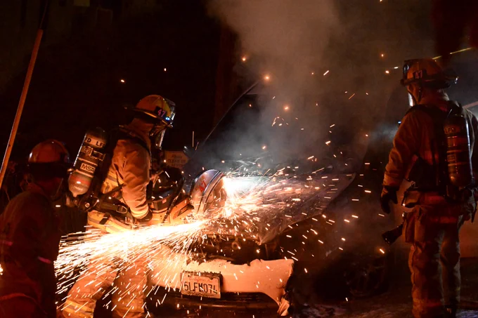 LAFD E47 cutting the hood of a vehicle on fire to gain access to the engine compartment.  This allows the fire department to put out the fire.  One firefighter cutting and another firefighter stands by as safety.