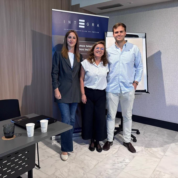 Tres personas (dos mujeres y un hombre) aparecen de pie en una sala de reuniones, delante de un roll-up de Integra Centros Especiales de Empleo. A la izquierda, una mesa con cuadernos, vasos de papel, un ordenador portátil y un teléfono móvil. Al fondo, una pared de madera y una pizarra tipo caballete.