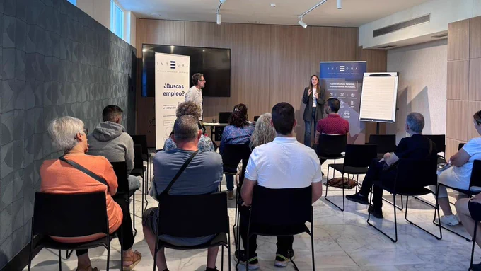 Grupo de personas sentadas en sillas dentro de una sala atienden hacia una presentación. Al frente, una persona interviene junto a dos roll-ups de Integra Centros Especiales de Empleo. En uno de ellos puede leerse: “¿Buscas empleo?”. A la derecha, una pizarra en blanco.