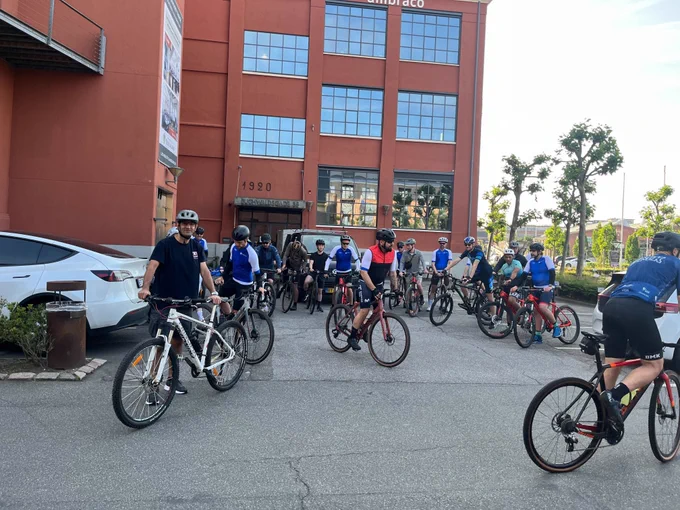 A group of cyclists gathered on their bicycles in front of a large red brick Umbraco office building.