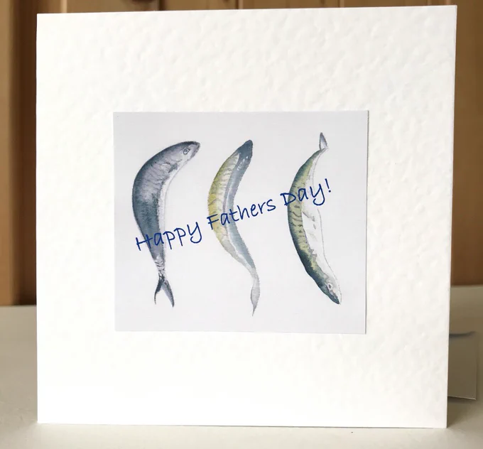 A white card with a blue and green watercolor illustration of three fish. The text 'Happy Fathers Day!' is written in blue cursive on the card.