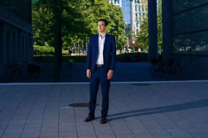 A man in a blue suit and glasses standing outdoors on a paved courtyard, featured in an article about the first generative AI worm.