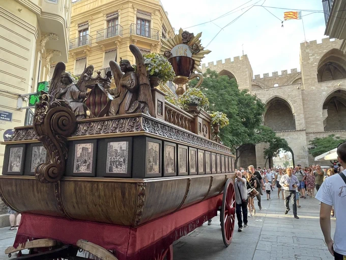 Carroza procesional tallada con ángeles, cáliz dorado y ramos, pasando junto a edificios históricos y una multitud.