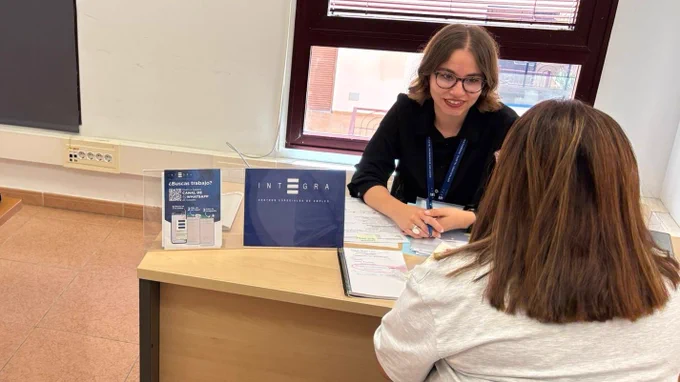 Dos personas sentadas frente a frente en una mesa mantienen una entrevista de trabajo en una sala interior. Sobre la mesa hay documentos, un cuaderno y material informativo de Integra CEE, incluyendo un cartel con código QR y el texto “¿Buscas trabajo? Canal de WhatsApp”. Al fondo, una ventana con persiana y, en la pared, un cartel informativo con un persona en silla de ruedas.