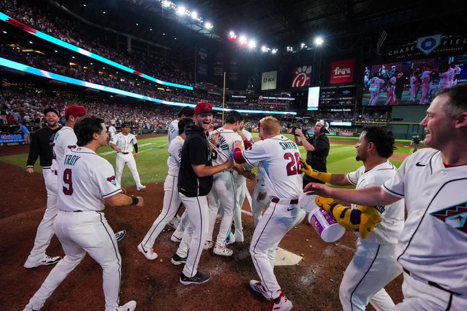 The team celebrates Ketel Marte at home plate after hitting a walk-off home run to win the game. They are wearing white Diamondbacks uniforms.