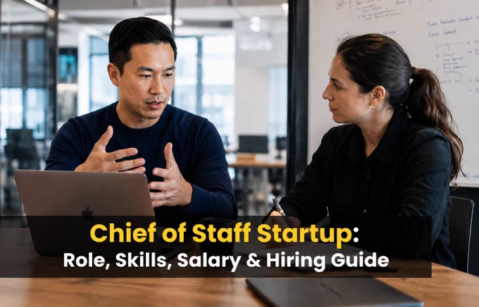 "Two professionals in a modern office are engaged in a focused business discussion. The man gestures as he explains a point, while the woman listens attentively and takes notes. A laptop and a whiteboard are visible in the background, with overlay text reading 'Chief of Staff Startup: Role, Skills, Salary & Hiring Guide'."