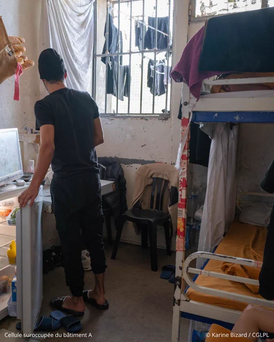 Photo d’une cellule vétuste de détention. Des draps blancs sont suspendus de chaque côté de la pièce. Un lit superposé est installé à droite. A gauche se trouve un lavabo et un miroir suspendu. Un détenu, debout de dos, ouvre le petit réfrigérateur sur lequel est posé une télévision. 
