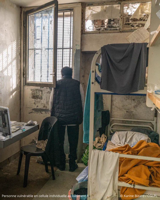 Photo d’une cellule individuelle de détention dont les murs sont usés et marqués. À droite de la pièce se trouve un lit superposé, un détenu est debout face à la fenêtre ouverte au fond de la cellule. À gauche sont installés un lavabo, un petit réfrigérateur surmonté d’un écran de télévision.