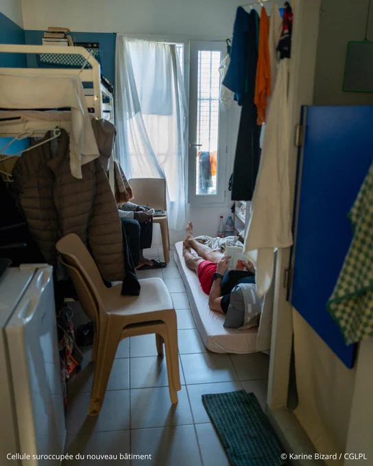 Photo d’une cellule suroccupée en détention. A gauche de la pièce sont installés un petit réfrigérateur et un lit superposé. A droite, on aperçoit la porte battante des sanitaires et du linge qui sèche. Un détenu est allongé sur un matelas par terre, en train de lire un livre. Un autre est assis sur le lit du bas. 