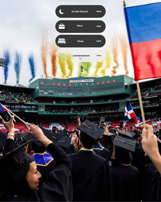 Graduates in caps & gowns celebrate commencement at Fenway Park, with colorful smoke over the stadium. Many hold phones; one has a Dominican Republic flag. 