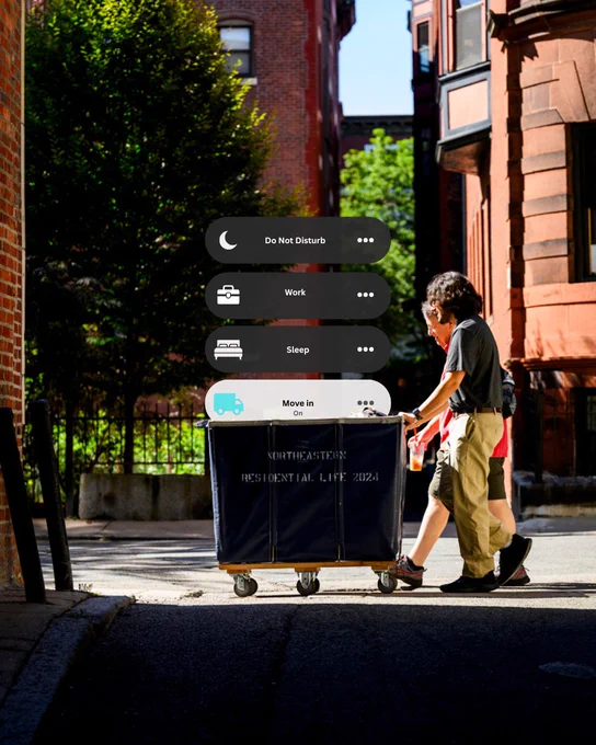 Two people push a blue laundry cart labeled 'NORTHEASTERN RESIDENTIAL LIFE 2024' down a sunny city street.