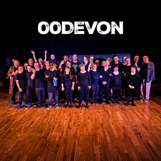 Large group of performers dressed mostly in black standing together on a theatre stage, smiling and waving for a group photo. A dark curtain backdrop and wooden stage floor frame the scene, with the word “OODEVON” displayed in large white letters above them.