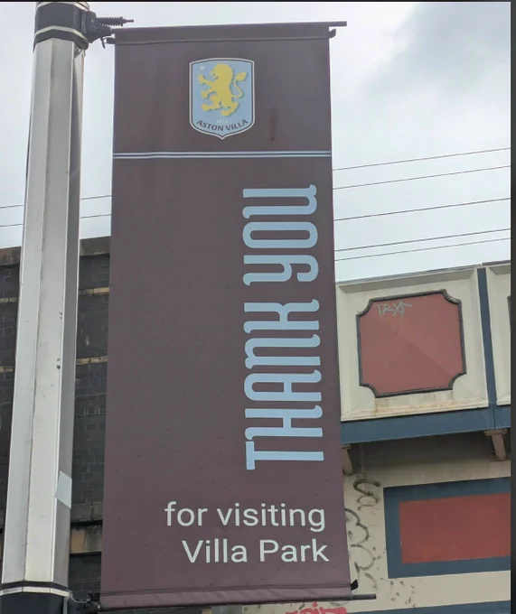 A street-level photograph of a tall, vertical fabric banner attached to a metal post. The banner is claret with light blue text. At the top is the Aston Villa F.C. crest, showing a yellow rampant lion on a light blue shield. Below a thin horizontal line, the words "THANK YOU" are printed vertically in a stylized, bold, light blue font. At the bottom, the phrase "for visiting Villa Park" is written horizontally in white. An urban building facade and an overcast sky are visible in the background.