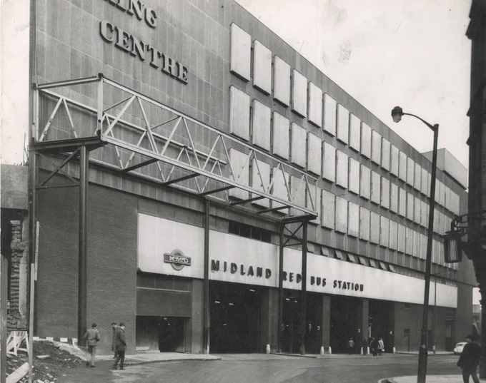 A photograph. The exterior of a 1960s building. Lettering in the top left of the photograph is clipped but should read Bull Ring Centre in its entirety. Three rows of uniform glass windows to the right. A metal gazebo structure in the bottom left of the photo. Facia below the window reads -  Midland Red Bus Station. Vehicle entrances below this. Pedestrians at various points on the street outside of the building. 