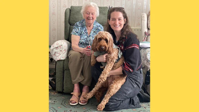 Josephine, 97, is sitting in a green armchair and Summer, 29, is kneeling in front of her to the left cuddling her dog