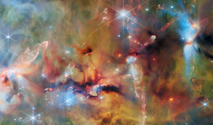 An area inside a star-forming molecular cloud. The background is covered with layers of gas and dust in blue, green and yellowish colours. Thicker clumps of cold dust, dark brown to black, block out light completely. Stars lie among and atop the clouds, from small orange ones to large white or blue ones. Waves and streams of glowing whitish gas are created by jets from protostars colliding with the surrounding material.
