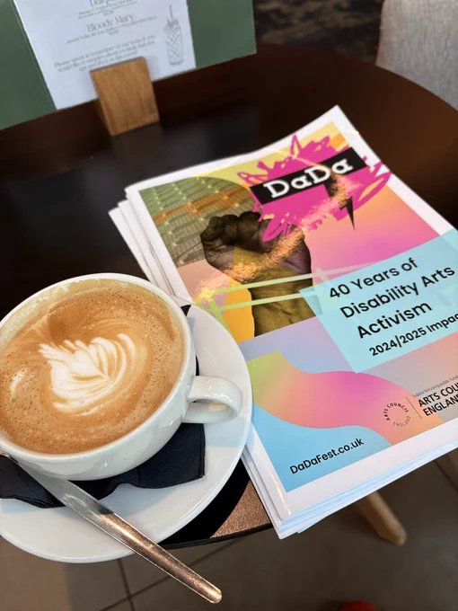A coffee cup sits on a table next to DaDa impact reports saying “40 years of disability arts activism”