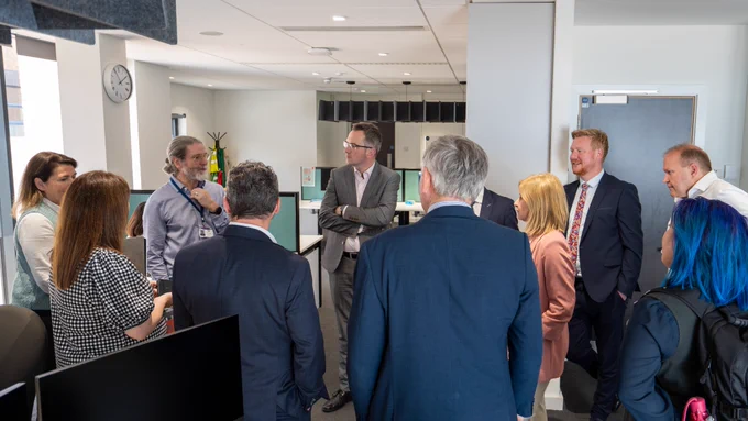 Group image showing members and staff from the committee listening to a man wearing a light coloured shirt. They are standing in an office space with light colour walls.