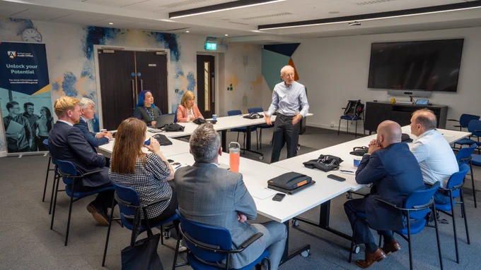 Eight people sit around table benches as a man wearing a light shirt and dark trousers stands in the middle leading a workshop session.